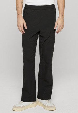 Mann iført svarte bukser med rette ben og glidelåslommer, hvite joggesko og en hvit skjorte, stående foran en ensfarget grå bakgrunn.