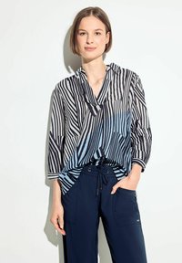 Blusa a righe bianche e nere con un gradiente blu, caratterizzata da un collo a V, maniche a trequarti e una vestibilità ampia, abbinata a pantaloni blu navy.