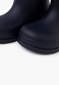 Gros plan sur des bottes de pluie en caoutchouc noir avec des semelles texturées sur fond blanc, mettant en valeur la zone des orteils et la partie inférieure de la tige.