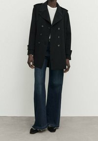 Czarny dwurzędowy płaszcz z dużymi klapami, w zestawie z szerokimi, ciemnymi dżinsami i czarnymi butami; gładka tekstura i dopasowany krój.