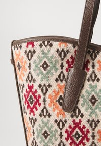 Woven fabric tote bag featuring a cream base with multicolored geometric patterns in red, green, orange, and black, complemented by brown leather handles.