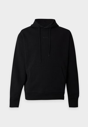 Jeune homme portant un sweat à capuche noir, un jean bleu large et des chaussures noires, debout devant un fond clair uni.