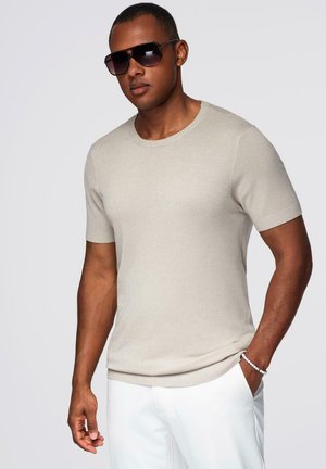 Beige short-sleeve sweater made of smooth knit fabric with a round neckline. Model wears dark sunglasses and white pants.