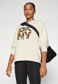 Beige kapucnis pulóver leopárdmintás NYC grafikával, laza fit, bordázott mandzsettákkal, fekete leggingsszel és egy textúrált fekete táskával párosítva.