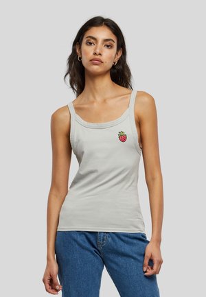 Femme portant un débardeur gris clair avec un petit motif fraise rouge sur la poitrine et un jean bleu sur fond gris uni.