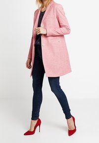 Cappotto in misto lana di un rosa chiaro, con colletto a punta, bottoni di chiusura singoli, tasche frontali, abbinato a jeans skinny blu scuro e tacchi alti rossi.