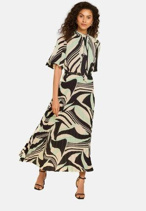 Femeie îmbrăcată într-o rochie maxi vaporoasă cu model, având dungi abstracte negre, bej și verde mentă, asortată cu pantofi cu toc și bretea la gleznă, negri.