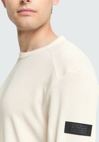 Lichtbeige longsleeve shirt met een gestructureerd wafelpatroon, ronde halslijn en zwart logo-patch op de linker mouw.