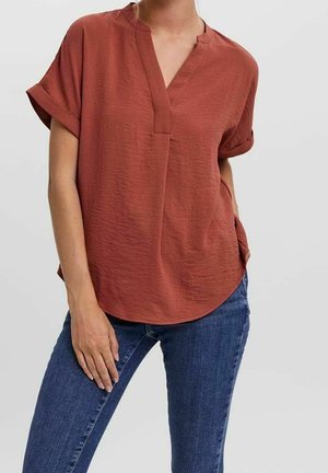 Vrouw draagt een roestkleurige blouse met korte mouwen, een V-hals en omslagen, gecombineerd met een blauwe spijkerbroek tegen een effen achtergrond.