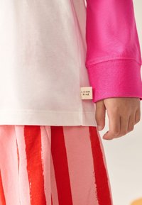 Valge t-särk roosade varrukatega ja väike „Little Bird“ silt. Sobitatud roosade pükstega, millel on punased vertikaalsed triibud. Pehme puuvillane materjal.