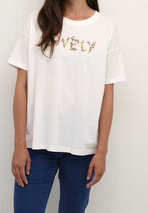 Kobieta w białej, oversize'owej koszulce z kwiatową wyszywanką "V E U", w niebieskich dżinsach, stojąca na jednolitym tle.