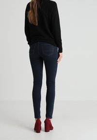 Úzké džíny v tmavě modrém denimovém provedení s kontrastním prošíváním, zadními kapsami, v kombinaci s burgundskými lodičkami na vysokém podpatku a černým svetrem.