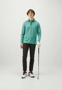 Μακρυμάνικο πόλο πουκάμισο σε ανοιχτό τιρκουάζ χρώμα με γιακά, μαύρο παντελόνι, λευκά παπούτσια γκολφ με κόκκινες λεπτομέρειες, κρατώντας ένα κλαμπ γκολφ.