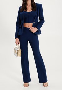 Žena v námořnickém blejzru, odpovídajících vysokých kalhotách a crop topu, držící metalickou zlatou kabelku a v otevřených lodičkách.