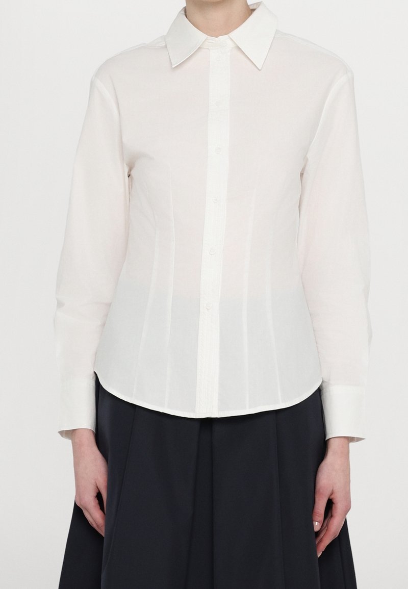 Femme portant une chemise blanche ajustée à manches longues avec un col classique, associée à une jupe noire.