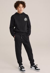 Melna džemperis ar logo, brīvs piegriezums, ribināti aproces. Saskaņots ar atbilstošām sporta biksēm ar kabatām. Nēsāts ar melniem un pelēkiem sporta apaviem.