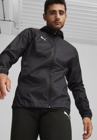 Schwarze leichte Jacke mit Reißverschluss und Kapuze. Ausgestattet mit elastischen Bündchen und einem weißen Logo auf der linken Brust. Glatte, glänzende Textur.