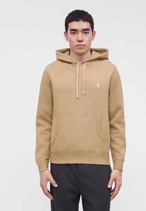 Jeune homme portant un sweat à capuche beige avec une poche avant et un petit logo blanc sur la poitrine, debout devant un fond uni.