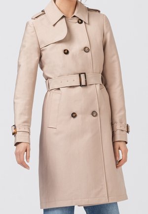 Trench-coat beige à double boutonnage avec ceinture, épaulettes et brides aux poignets, porté sur un jean bleu.