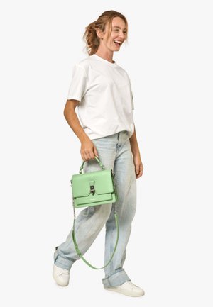 Femme souriante marchant, portant un t-shirt blanc, un jean bleu clair, des baskets blanches et tenant un sac à main vert par sa anse supérieure.