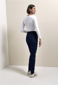 Witte top met lange mouwen gecombineerd met donkerblauwe skinny jeans, met een aansluitende pasvorm. Sneakers hebben een logo met groene en rode accenten.