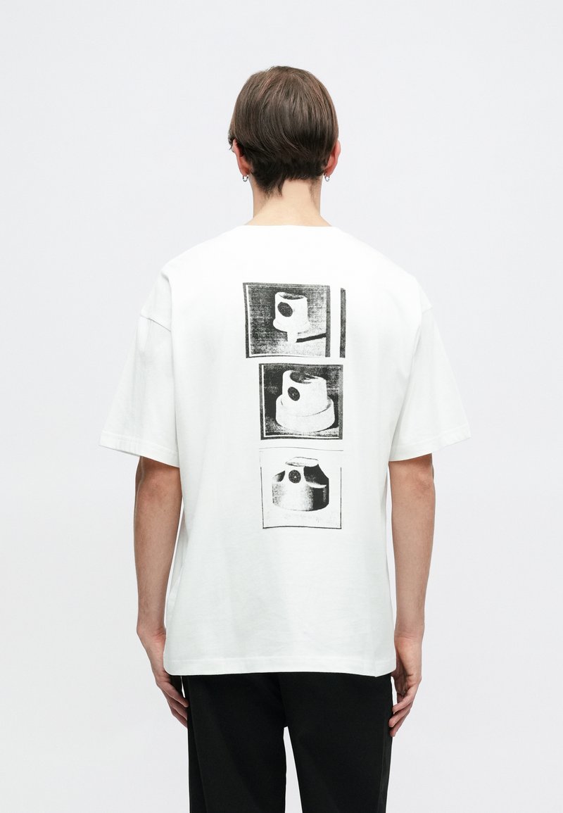 Camiseta blanca de algodón con tres impresiones gráficas negras en la parte posterior. Corte holgado con manga corta y cuello redondo.