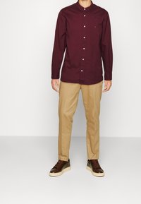 Camisa marrón con cuello mandarín, acompañada de pantalones caqui. El conjunto se completa con zapatillas deportivas marrón oscuro.