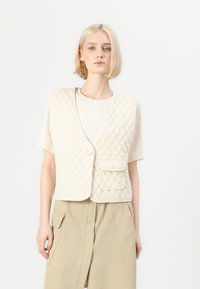Quilted beige vest med v-hals, med knappelukking og en frontlomme. Brukt over en plissert lys topp og en beige skjørt.