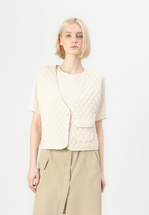Quilted beige vest med v-hals, med knappelukking og en frontlomme. Brukt over en plissert lys topp og en beige skjørt.