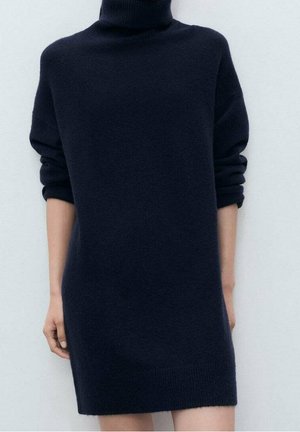 Personne portant une robe-pull à col roulé bleu marine à manches longues, manches retroussées jusqu'aux avant-bras, debout devant un fond uni.