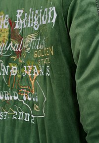 Groene, gestructureerde stoffen jas met levendige geborduurde ontwerpen in het wit, geel en rood, waaronder een arend, bomen en tekst.