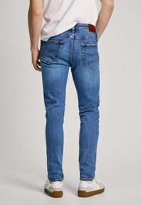Jeans de denim azul ajustados con costuras visibles, dos bolsillos traseros y una etiqueta de cuero marrón en la cintura, combinados con zapatillas blancas.
