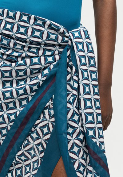 Άτομο που φοράει μπλε μπλούζα και περιτυλιγμένη φούστα με γεωμετρικό μοτίβο, δεμένη στη μέση, με σκίσιμο στο πόδι και ακάλυπτο χέρι.