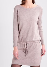 Knitklänning i ljusbeige med långa ärmar, ribbad struktur och dragsko i midjan. Har rund halsringning och en avslappnad passform.