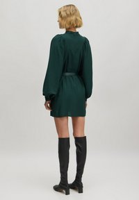 Dunkelgrünes Mini-Kleid mit Rüschensleeves, gerafften Details an den Schultern und einem Stoffgürtel. Kombiniert mit knielangen schwarzen Lederstiefeln.
