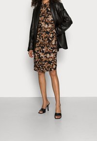 Svart läderblazer över en mönstrad svart klänning med orange och vita blommönster. Kvinnan bär svarta klackskor. Släta texturer.