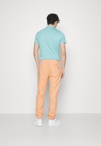 Mann, der mit dem Rücken zur Kamera steht, trägt ein hellblaues Poloshirt, Pfirsichfarbene Jogginghosen und weiße Turnschuhe vor einem schlichten weißen Hintergrund.