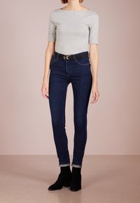 Osoba v šedém tričku s krátkým rukávem, tmavě modrých skinny džínách s černým pásem a černých kotníkových botách stojící na béžové podlaze.