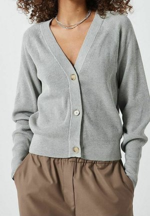 Lys grå ribbet cardigan med V-hals, fem knapper og lange ærmer. Båret over brune bukser med tekstureret materiale og tætsiddende design.