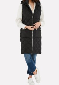 Femme portant un long gilet sans manches noir matelassé avec fermeture éclair, un chemisier blanc, un jean bleu raccourci et des baskets blanches, debout les jambes croisées.