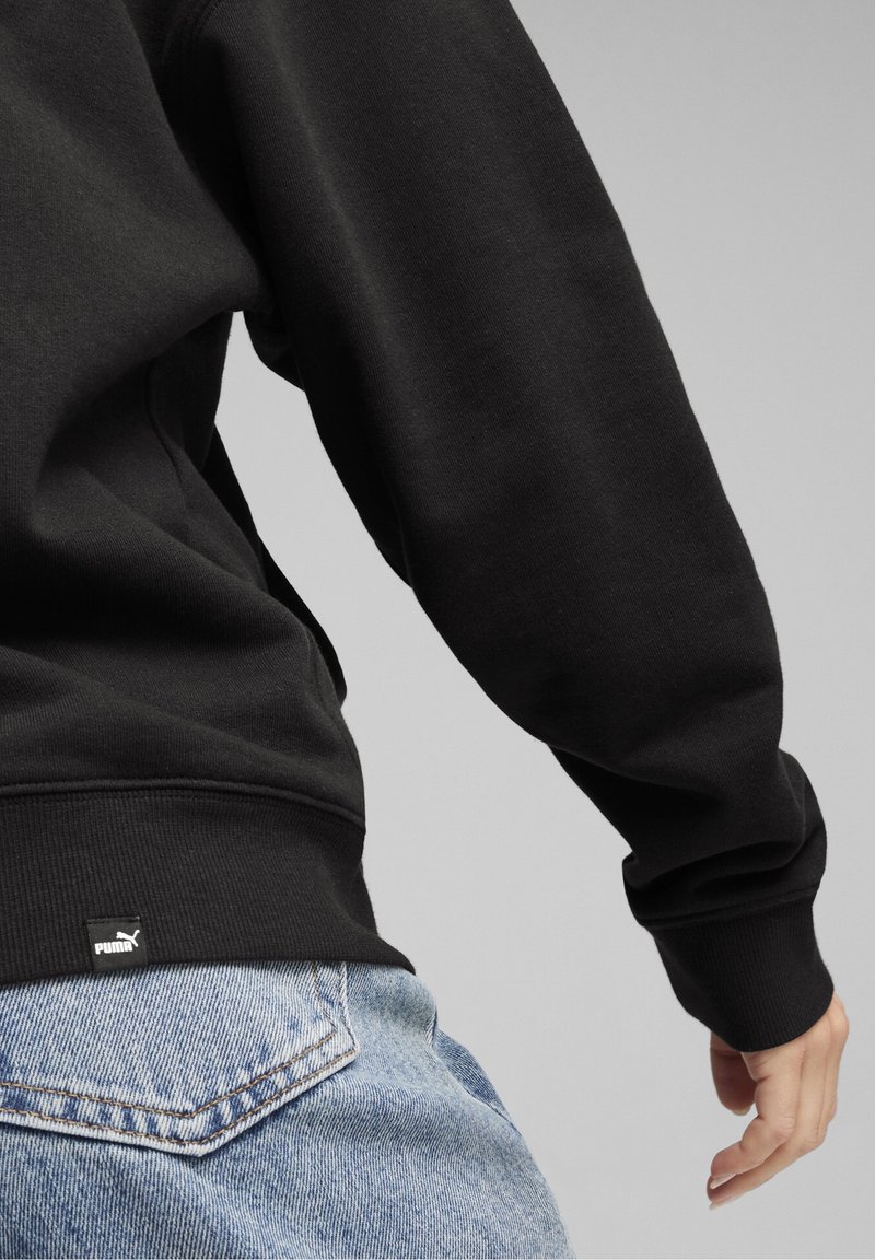 Personne portant un sweat-shirt Puma noir et un jean bleu clair, montrant le bas du dos et le bras droit sur un fond gris uni.