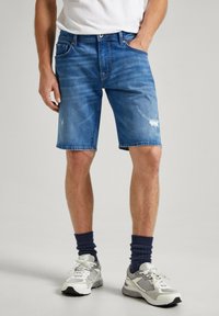 Shorts de mezclilla de color azul claro con un corte relajado, que presentan desgaste y detalles rasgados. Combinados con una camiseta blanca y zapatillas grises.