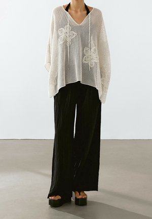 Femme portant un poncho blanc en crochet avec motifs floraux, pantalon noir à jambes larges et sandales noires à plateforme, debout sur un sol gris.