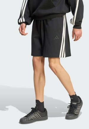 Person iført sorte Adidas-shorts, sorte sneakers og sorte sokker med tre hvide striber på siderne og synligt logo.
