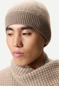 Beige stickad mössa med strukturerat mönster och ribbad nederkant; mjukt tyg. Matchas med en beige halsduk med liknande textur. Närbild.