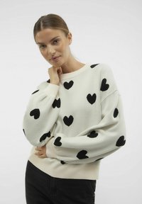 Pull blanc avec de grands motifs en forme de cœur noir, poignets et ourlet côtelés. Texture douce avec des manches légèrement bouffantes.
