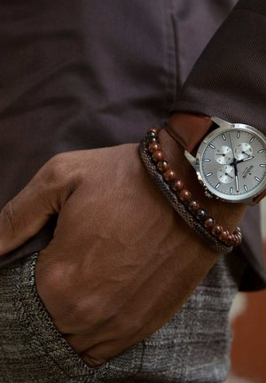 Zilveren horloge met drie sub-dials, bruine leren band en een geweven armband met donkere en roodachtige kralen, gedragen op een gestructureerde grijze stof.