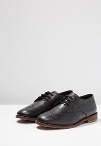 Černé kožené šněrovací boty s kulatou špičkou, brogue detaily a nízkým dřevěným podpatkem. Hladký povrch s perforovanými akcenty.