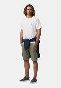 T-shirt bianco con logo blu, pantaloni cargo oliva con toppe e un maglione blu legato in vita. Scarpe verdi completano l'outfit.