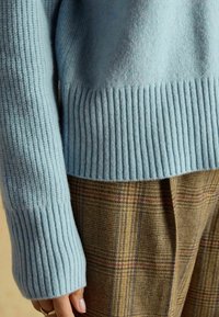 Pulover tricotat bleu deschis, cu manșete și tiv cu nervuri, asortat cu pantaloni în carouri ciocolatii, având mai multe nuanțe și un finisaj texturat.